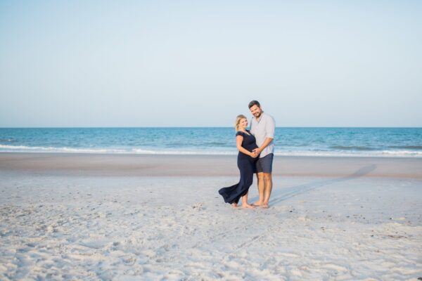 A couple stands on a sandy beach in front of the ocean, smiling and holding each other. The woman is visibly pregnant, wearing a long navy dress, while the man wears a light shirt and shorts. The sky is clear and blue.