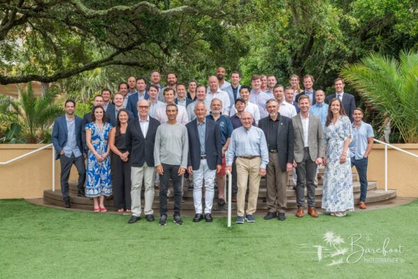 A large group of people, dressed in business casual and formal attire, pose together outdoors on green grass with trees and plants in the background, smiling at the camera.