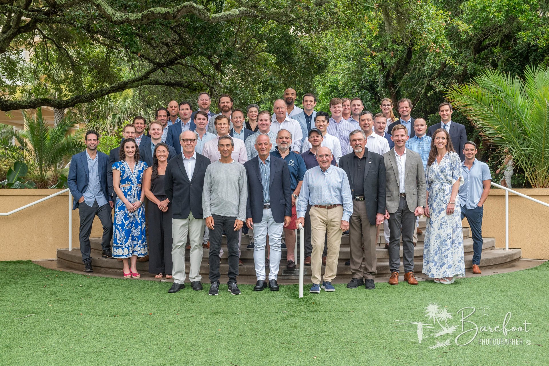 A large group of people, dressed in business casual and formal attire, pose together outdoors on green grass with trees and plants in the background, smiling at the camera.