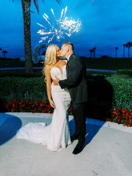 A bride and groom kiss outdoors at night in front of a garden, with fireworks lighting up the blue sky behind them. The bride wears a strapless white gown and the groom is in a black suit.