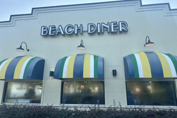 The exterior of Beach Diner with three striped awnings in yellow, green, black, and white, and three wall lamps above large foggy windows.