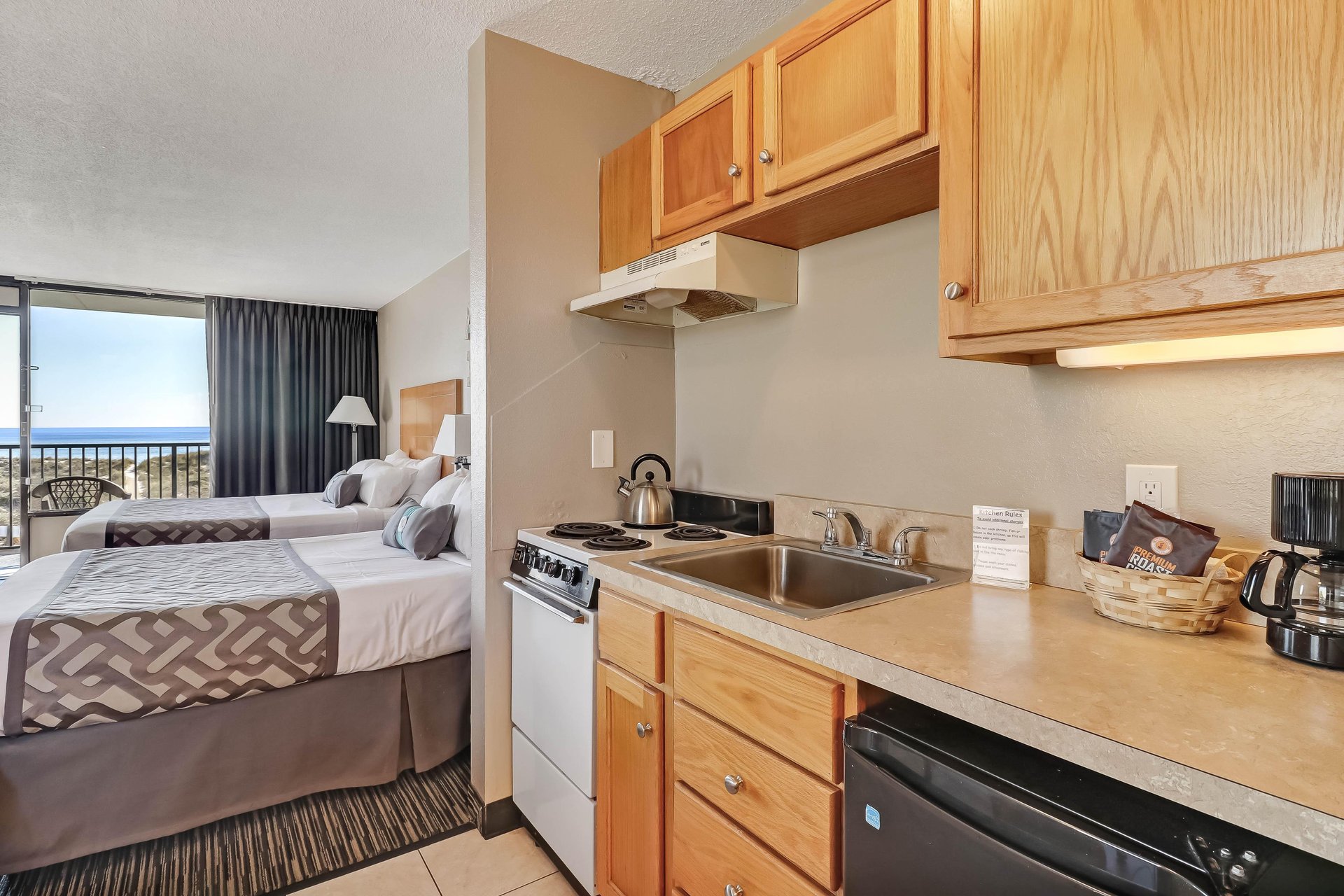 Hotel room with two double beds, a small kitchenette featuring a sink, stove, mini fridge, coffee maker, and a basket, plus a balcony with a view through sliding glass doors.