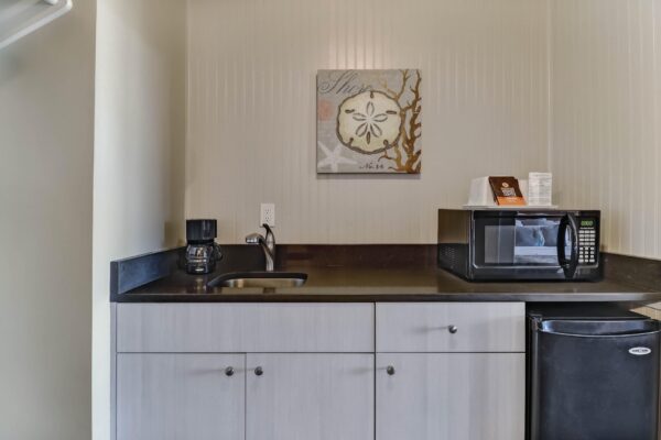 A small kitchenette with a sink, coffee maker, microwave, mini fridge, and wall art above the counter in a neutral-toned room.