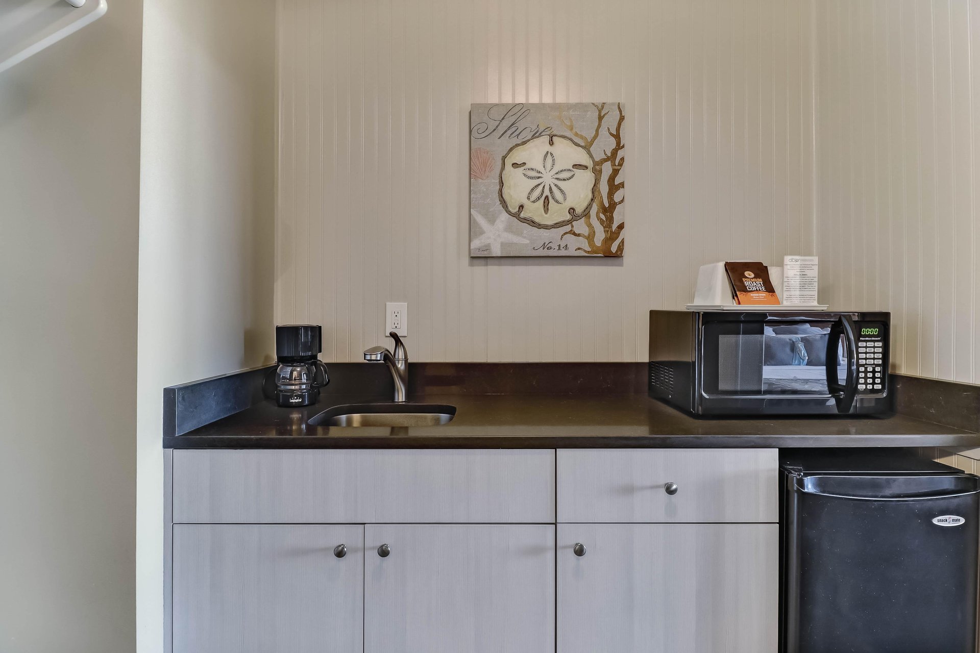 A small kitchenette with a sink, coffee maker, microwave, mini fridge, and wall art above the counter in a neutral-toned room.