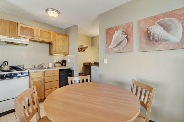 Small kitchen with wooden cabinets, a stove, sink, kettle, and dishwasher. A round wooden table with four chairs is in the foreground. Two shell-themed pictures hang on the beige wall above the table.