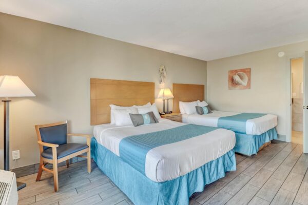 Hotel room with two double beds, blue and white bedding, pillows, a wooden chair, lamp, nightstand, wall art, and tiled floors. Door to bathroom is open on the right side of the image.
