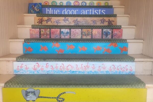 Colorful painted stair risers decorated with artwork featuring a cat, fish, flamingos, spirals, owls, turtles, and the words blue door artists at the top step.