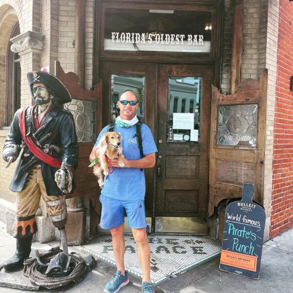 A man in a blue outfit holding a small dog stands in front of Floridas Oldest Bar next to a pirate statue and a sign advertising World Famous Pirates Punch. The entrance is flanked by wooden doors.