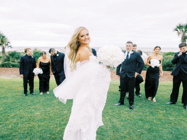 A smiling bride in a white dress holds a white bouquet, standing on grass with bridesmaids and groomsmen in black outfits behind her. The outdoor setting includes greenery and a cloudy sky.