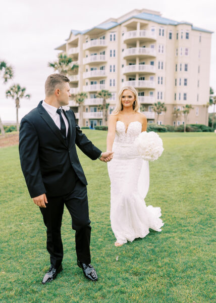 A bride in a strapless white gown holds a bouquet and walks hand in hand with a groom in a black suit on green grass, with a multi-story building and palm trees in the background.