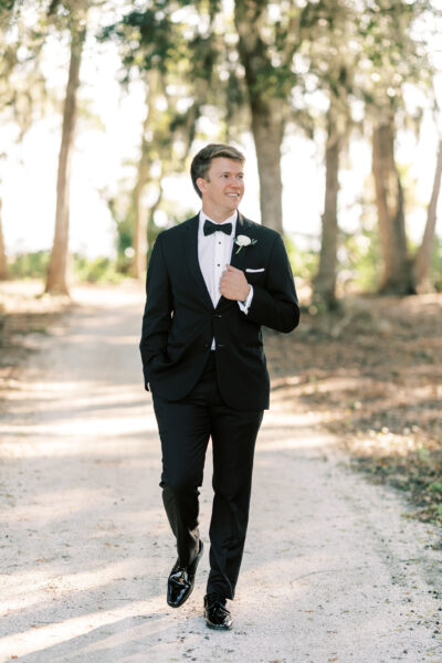 A man in a black tuxedo with a bow tie and boutonnière walks along a sunlit, tree-lined path, smiling and looking to the side with one hand in his pocket.