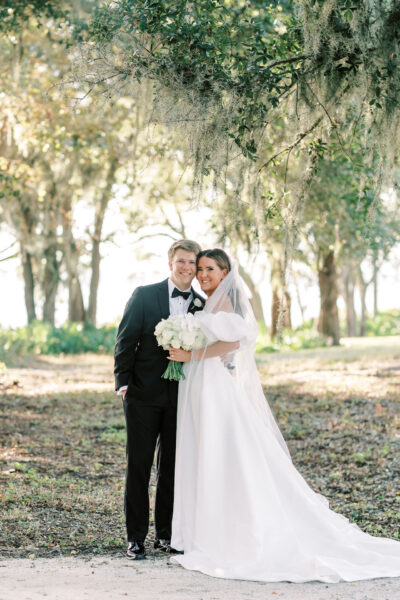 A bride in a white gown and veil stands beside a groom in a black tuxedo. They smile and embrace outdoors under trees, with sunlight filtering through the leaves. The bride holds a bouquet of white flowers.