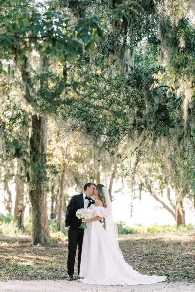 A bride and groom kiss under tall, moss-draped trees in a sunlit, outdoor setting. The bride wears a white gown and veil, holding a bouquet of white flowers, while the groom is dressed in a black tuxedo.