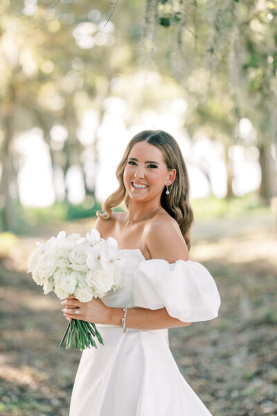 A bride in an off-the-shoulder white gown smiles while holding a bouquet of white flowers. She stands outdoors in a sunlit, wooded area with soft, greenery in the background.