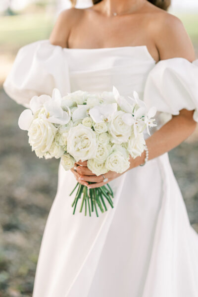 A bride in an off-the-shoulder white dress holds a bouquet of white roses and orchids. Her hands display a ring and bracelet. The background is softly blurred.