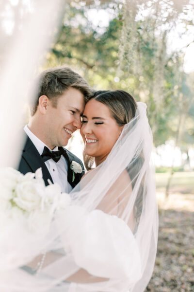 A bride and groom stand close together outdoors, smiling joyfully under the bride’s veil. The groom wears a black tuxedo and boutonnière, while the bride holds a bouquet of white flowers. Sunlight filters through trees in the background.