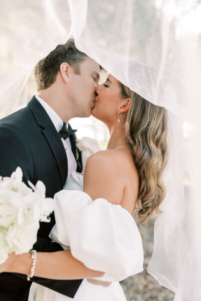 A bride and groom share a kiss under a sheer white veil. The bride holds a bouquet of white flowers and wears an off-the-shoulder dress, while the groom is in a black tuxedo. Soft lighting creates a romantic atmosphere.