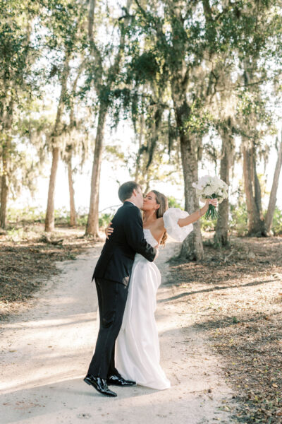 A bride in a white dress holds a bouquet and smiles as she is kissed on the cheek by a groom in a black suit on a sunlit forest path.