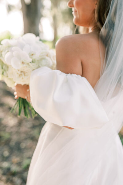 A bride in an off-the-shoulder white wedding dress holds a bouquet of white flowers, with sunlight softly illuminating her and a blurred natural background.