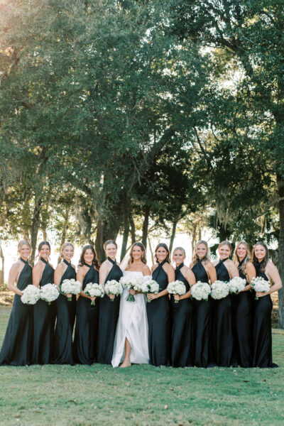 A bride in a white dress stands outdoors with eleven bridesmaids in matching black dresses, all holding white bouquets and smiling, with trees in the background.