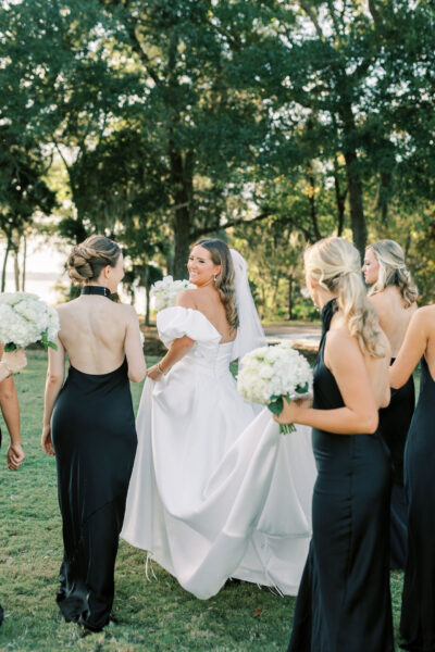 A bride in a white gown and veil smiles while walking outside with four bridesmaids in black dresses, all holding white floral bouquets. Sunlight filters through trees in the background.