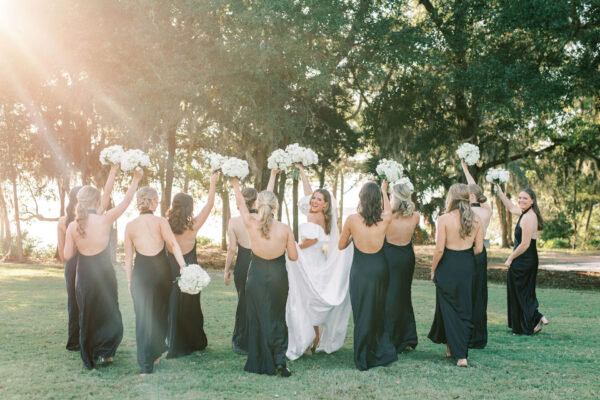 A bride in white is surrounded by bridesmaids in black dresses, all raising white bouquets in an outdoor, sunlit setting with trees and grass.