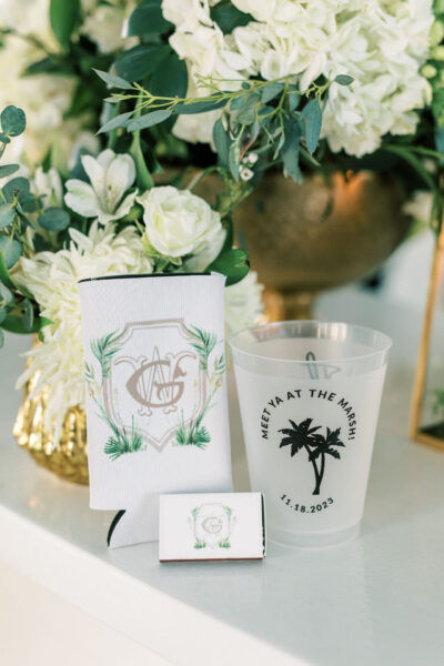 A close-up of wedding favors includes a monogrammed koozie, a clear cup with palm trees and “Meet Ya At The Marsh 11.18.2023,” and a matchbox, all set in front of a white and green floral arrangement.