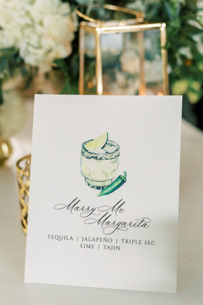 A card with an illustration of a margarita garnished with lime and a jalapeño. Text reads: Marry Me Margarita. Tequila | Jalapeno | Triple Sec | Lime | Tajin. Greenery and a candle are in the background.