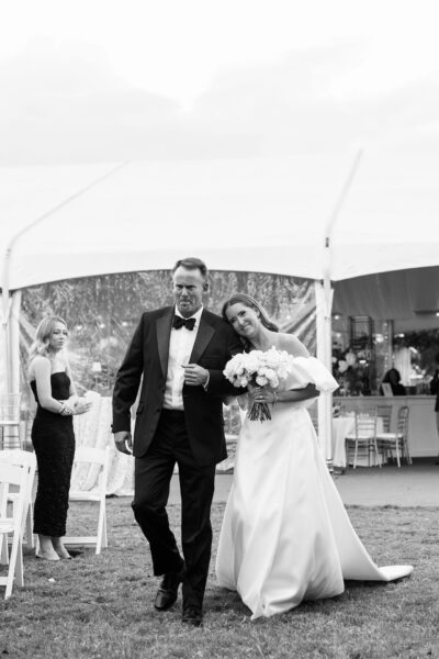 A bride in a white gown holding a bouquet walks arm-in-arm with a man in a tuxedo. She smiles and leans her head on his shoulder. A bridesmaid stands in the background. They are outside near a white tent.