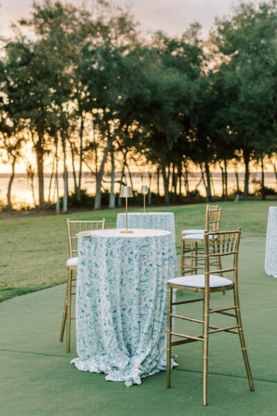 Tall cocktail tables with patterned tablecloths and gold chairs are set up outdoors on green grass, with trees and a lake visible in the background at sunset.