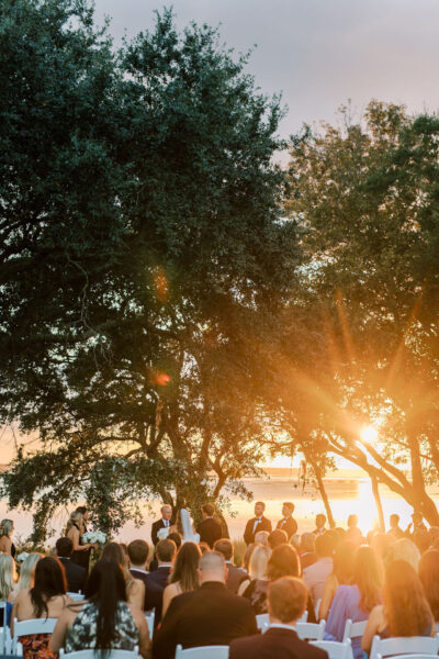 An outdoor wedding ceremony takes place at sunset beneath large trees, with guests seated and sunlight streaming through the branches, creating a warm, golden glow.