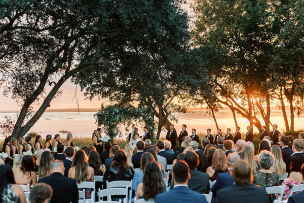 A wedding ceremony takes place outdoors at sunset near a waterfront, with guests seated facing a couple and officiant under large trees. The wedding party stands on either side, bathed in warm, golden light.