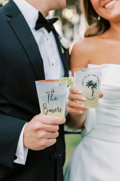 A bride and groom dressed in formal attire toast with personalized plastic cups; the bride’s cup has a palm tree design and date, and the groom’s cup reads “The Busers,” both holding cocktails garnished with lime and chili salt rims.