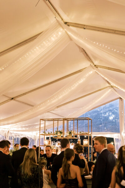 Guests in formal attire gather under a white tent with draped fabric and string lights, socializing near a bar. A mountain is visible through the clear tent walls in the background.