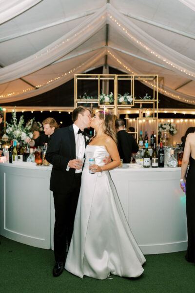 A bride and groom in formal attire share a kiss in front of a white bar decorated with flowers and bottles, under a tent with draped fabric and string lights at a wedding reception.