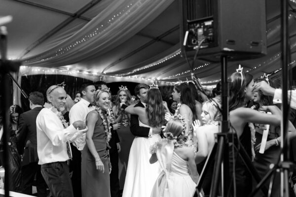 A joyful group of people dance and celebrate together at a wedding reception under a tent decorated with string lights. The bride and groom hug at the center, surrounded by smiling guests. The image is in black and white.