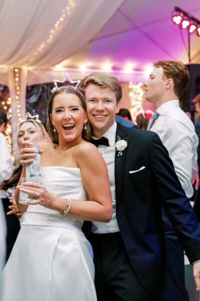 A smiling bride in a white dress and a groom in a black tuxedo pose together on a lively dance floor, surrounded by guests under colorful lights at a wedding reception.
