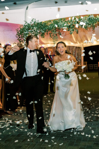 A bride in a white gown holding a bouquet and a groom in a black tuxedo walk joyfully together under falling flower petals, surrounded by cheering guests at a festive, outdoor evening celebration.