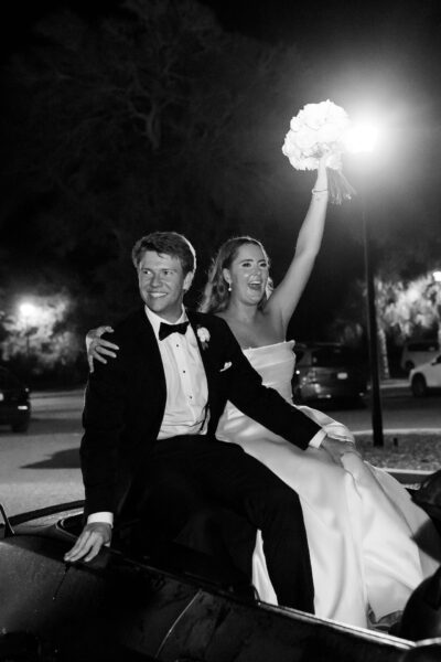 A bride in a strapless gown holds a bouquet high and smiles joyfully while sitting beside her smiling groom in a tuxedo; both are seated on a car at night.