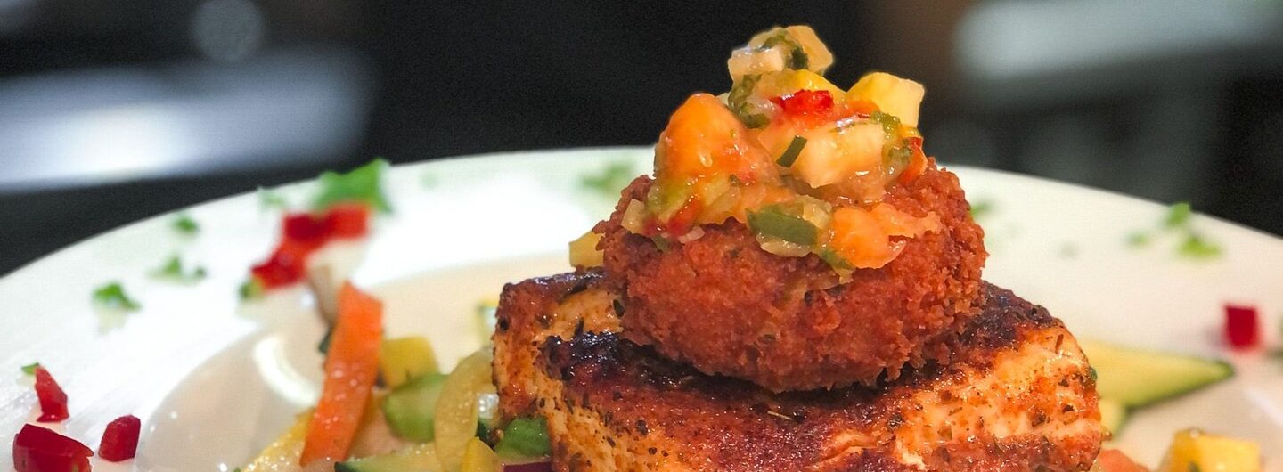 A white plate with grilled fish and a golden-fried seafood cake on top, served over white rice and sautéed vegetables, garnished with herbs and diced peppers. A blurred person stands in the background.