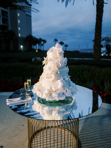 A tall white wedding cake decorated with cascading white flowers sits on a mirrored round table beside two champagne glasses, set outdoors in the evening near palm trees and a building.