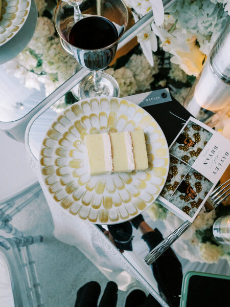 A slice of layered cake sits on a decorative plate next to a glass of red wine, a fork, and a printed event menu atop a reflective table decorated with white flowers.