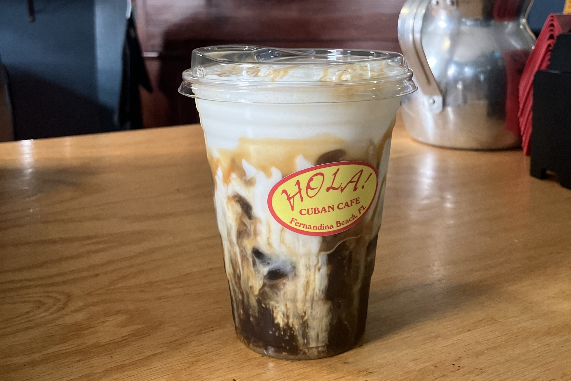 A plastic cup of iced coffee with cream swirls sits on a wooden table. The cup has a clear lid and a Hola Cuban Cafe sticker on it. The background shows a silver pitcher and red napkins.
