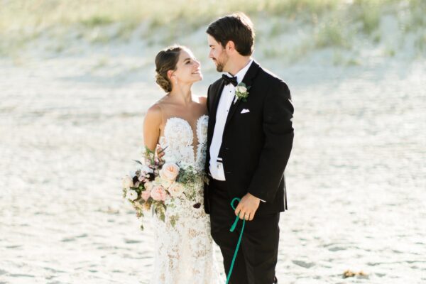 A bride in a lace gown holding a bouquet and a groom in a tuxedo stand on a sandy beach, smiling and gazing at each other lovingly.