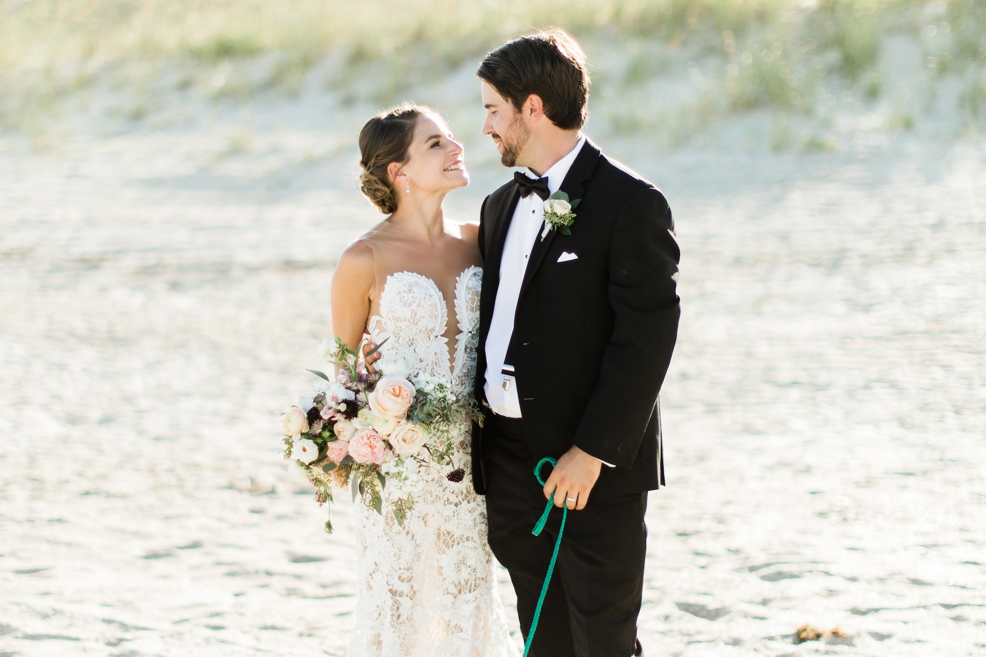 A bride in a lace gown holding a bouquet and a groom in a tuxedo stand on a sandy beach, smiling and gazing at each other lovingly.
