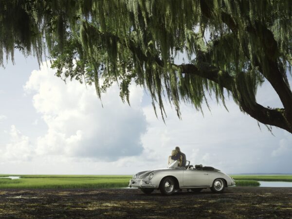 A person sits in a vintage white convertible parked under a large tree with hanging Spanish moss, overlooking a serene landscape of green fields and water under a bright sky with scattered clouds.
