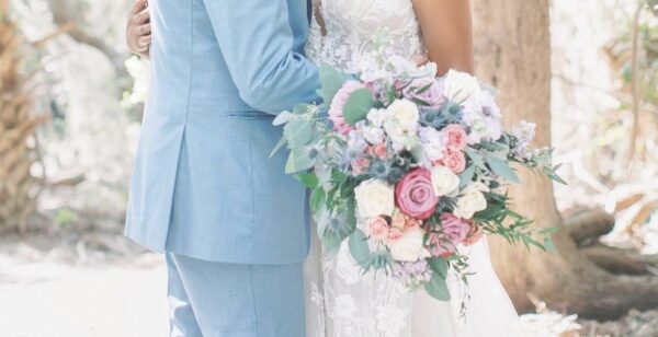 A couple dressed in wedding attire embrace outdoors. The bride holds a large bouquet of pink, purple, and white flowers with greenery. Sunlight filters through trees in the background.