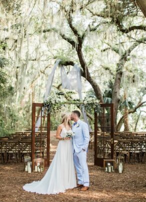 A bride and groom kiss at an outdoor wedding ceremony under trees with draped fabric, wooden chairs, and lanterns. The setting is romantic and rustic with greenery and natural light.