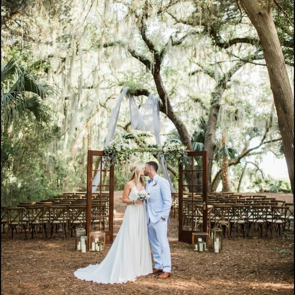 A bride and groom kiss at an outdoor wedding ceremony under trees with draped fabric, wooden chairs, and lanterns. The setting is romantic and rustic with greenery and natural light.