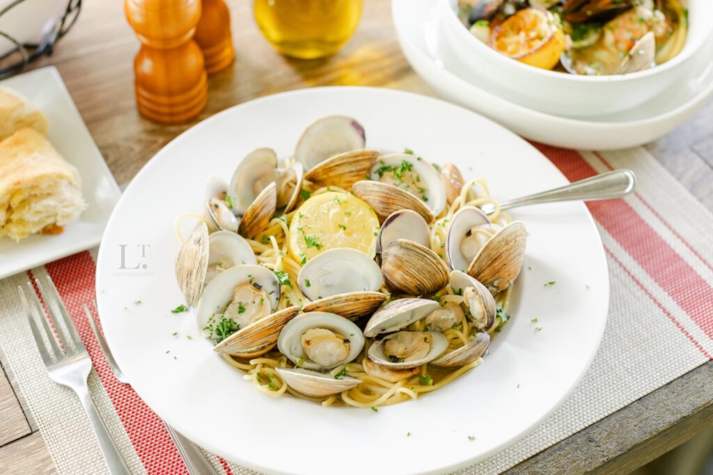 A plate of spaghetti topped with clams, lemon slices, and chopped herbs sits on a table next to bread, a pepper shaker, and a bowl of seafood, with a fork resting on the plate.